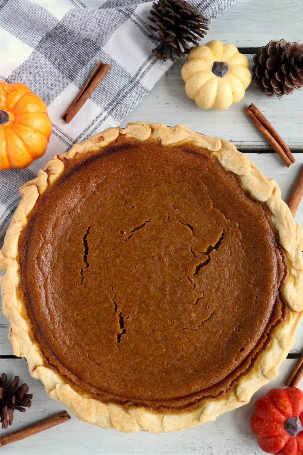 A full pumpkin pie surrounded by pine cones, pumpkins, and cinnamon sticks.