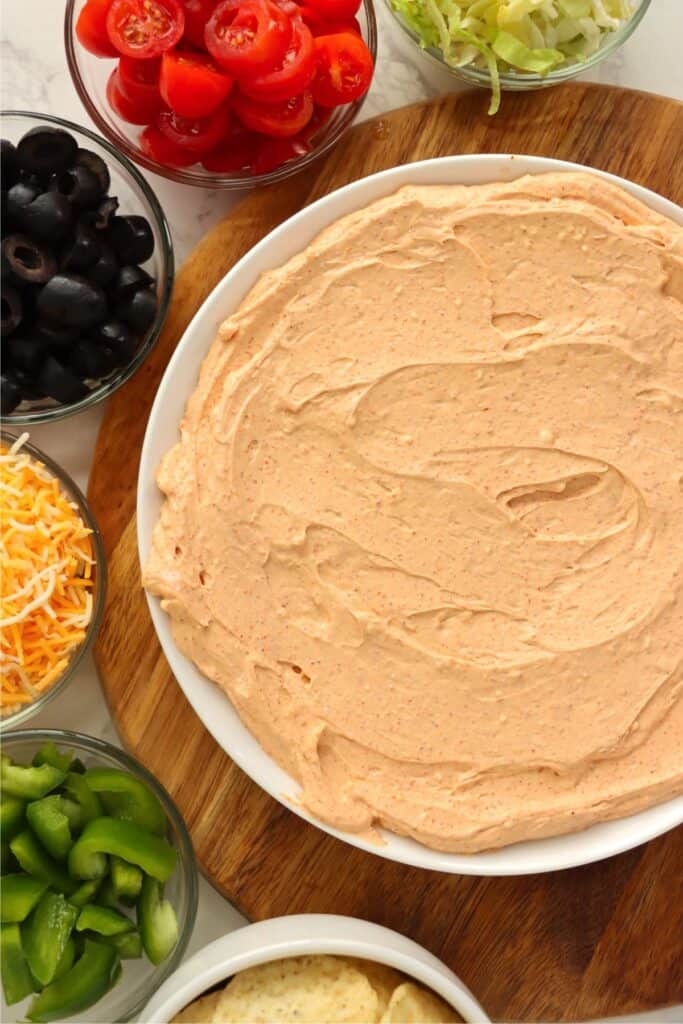 Overhead shot of bowlful of fiesta taco dip surrounded by bowls of olives, bell peppers, tomatoes, and cheese. 